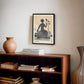 Black-framed Lady Justice artwork displayed above a wooden bookcase with stacked books and a ceramic vase.