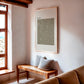 Framed artwork above a wooden bench with pillows in a room with large windows.
