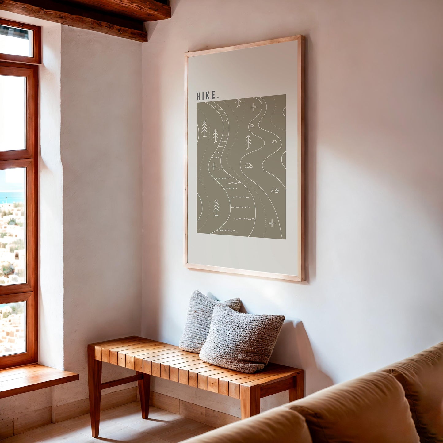 Framed artwork above a wooden bench with pillows in a room with large windows.