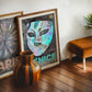 Dark wood-framed Venice carnival mask artwork leaning with a Paris print behind a striped wooden vase.