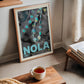 Oak-framed NOLA 1857 Mardi Gras bead artwork leaning beside a window in a living room with books and a cup of tea.