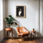 Oak-framed The Stylist art print displayed above a tan leather chair between two potted plants.