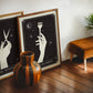 Dark wood-framed The Colorist artwork leaning beside The Stylist artwork, a striped wooden vase, plant, and brown stool.