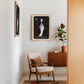 Oak-framed The Colorist artwork displayed above an armchair beside a potted plant and wooden cabinet.