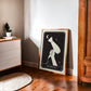 Oak-framed Blowout art print leaning against a wall between a dresser and wooden wardrobe.