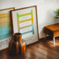 Wood-framed Polyvagal Ladder diagram leaning against a wall beside another colorful chart, a striped vase, and a brown stool.
