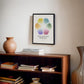 Black-framed Feelings Wheel art print hanging above a wooden bookcase with stacked books and a ceramic vase.