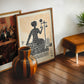 Oak-framed Lady Justice artwork displayed in an interior setting beside another framed print, a striped wooden vase, and a brown stool.