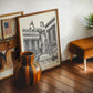 Wood-framed black-and-white Blind Justice artwork leaning beside courtroom art, a striped wooden vase, and a brown stool.