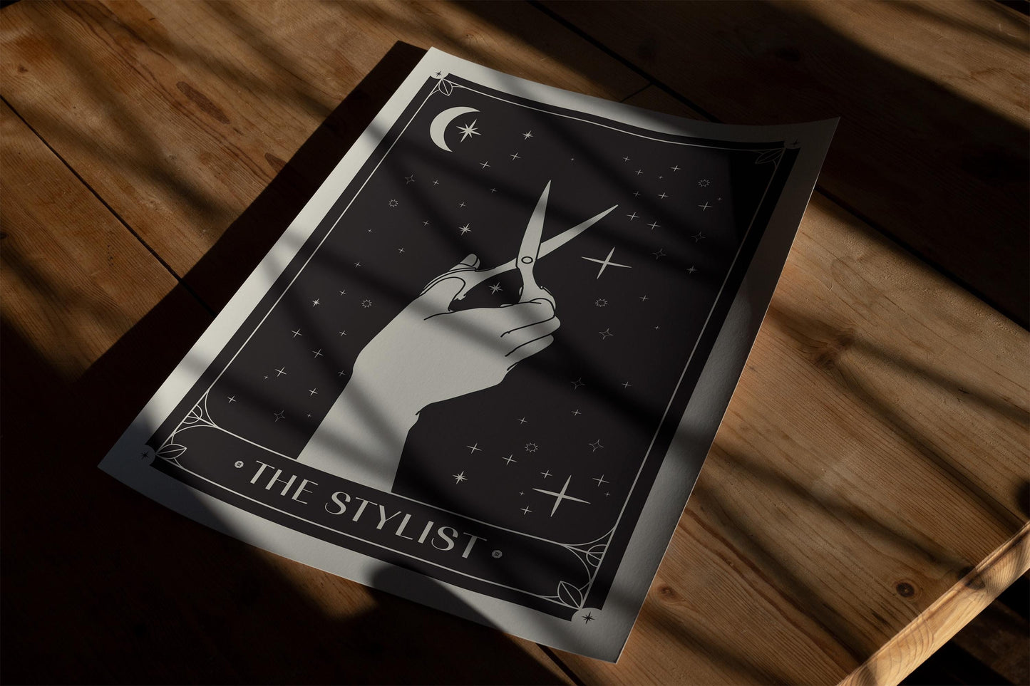 Unframed The Stylist art print showing a hand holding scissors, resting on a wooden table in striped sunlight.
