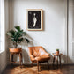Oak-framed The Colorist artwork displayed above a tan leather armchair between two potted plants.