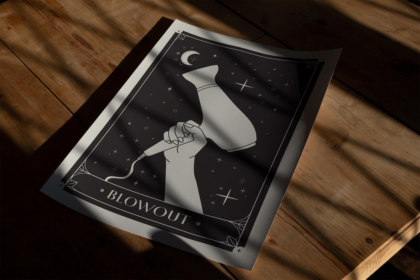 Unframed Blowout art print depicting a hand holding a hair dryer beneath a crescent moon and stars, laid on a wooden surface.