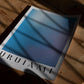 Unframed Silent Reflection No.1 blue and lavender gradient art print lying on a wooden surface beneath leafy shadows.