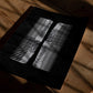 Unframed black art print with two white comb-like forms lying on a wooden table in patterned sunlight.
