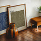 Dark wood-framed HIKE. artwork leaning beside a blue bicycle print, striped vase, and tan stool.