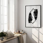 Black-framed abstract black-and-white artwork displayed in a sunlit interior beside tall windows and plants.