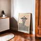 Oak-framed Caduceus art print leaning on a wood floor beside a cabinet and potted plant.