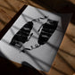 Unframed abstract black-and-white art print with two textured forms and curling lines on a sunlit wooden table.