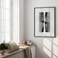 Black-framed abstract comb artwork displayed in a sunlit interior beside tall windows and plants.