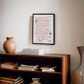 Black-framed art print with red handwritten dining advice displayed above a wooden bookcase with books and a ceramic vase.