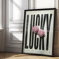 Black-framed Lucky artwork with oversized black lettering and pink fuzzy dice, leaning against an ornate white paneled wall.