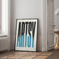 Black-framed Artsy typography artwork with elongated black-to-blue letters, leaning beside a window in a bright interior.