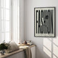 Black-framed FASHION typography artwork with button and thread displayed in a sunlit room.
