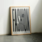 Oak-framed FASHION typography artwork with button and thread leaning against a sunlit wall.