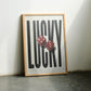 Oak-framed LUCKY artwork with two red dice leaning against a sunlit wall between chairs.