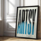 Black-framed Artsy typography artwork with elongated black-to-blue letters, leaning against a sunlit paneled wall.