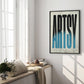 Black-framed Artsy typography artwork displayed on a bright wall beside tall windows, books, cushions, and greenery.