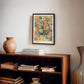 Black-framed Cerebrum in Bloom art print displayed above a wooden bookcase with books and a ceramic vase.
