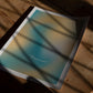 Unframed teal and yellow abstract “Get back to center” art print lying on a wooden table in patterned sunlight.