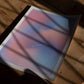 Unframed pink and blue gradient art print reading “The only way out is through” lying on a wooden table beneath window shadows.
