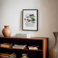 Black-framed 1965 Porsche 911 collage artwork displayed above a wooden bookcase with books and a ceramic vase.