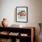 Black-framed orange BMW 2002 artwork hanging above a wooden bookcase with books and ceramic decor.