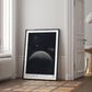 Black-framed Artemis II lunar horizon art print leaning against the wall in a bright room beside a window and open doorway.