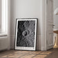 Black-framed Artemis II lunar crater art print leaning against a wall in a bright room beside a window and open doorway.