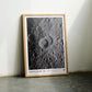 Oak-framed Artemis II lunar crater art print leaning against a pale wall between green and brown chairs.