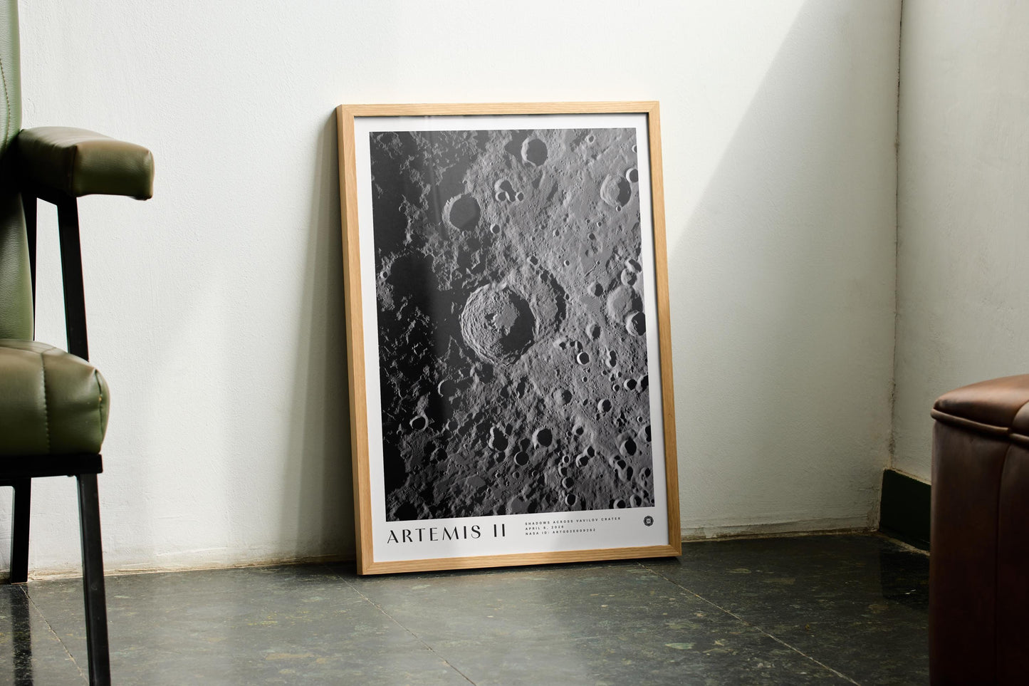 Oak-framed Artemis II lunar crater art print leaning against a pale wall between green and brown chairs.