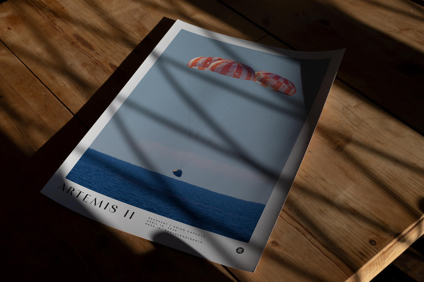 Unframed Artemis II art print showing the Orion capsule descending beneath three parachutes, lying on a wooden table in patterned sunlight.