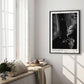 Black-framed Artemis II astronaut art print displayed on the wall of a sunlit room beside tall windows and a cushioned bench.