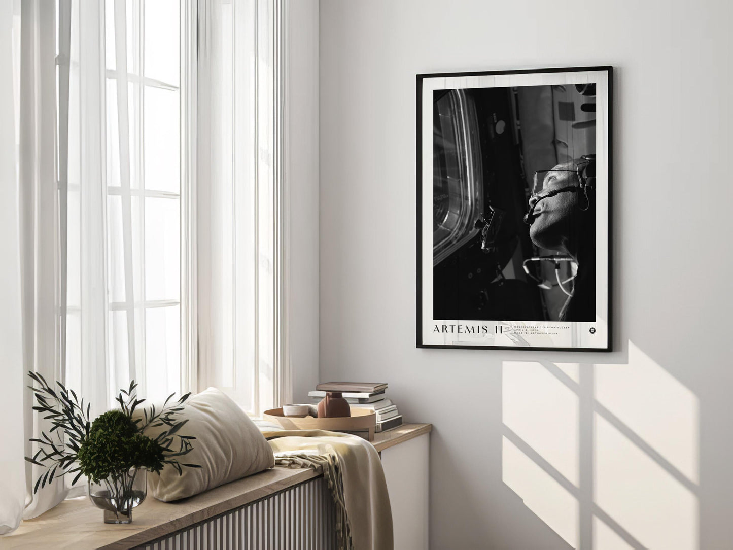Black-framed Artemis II astronaut art print displayed on the wall of a sunlit room beside tall windows and a cushioned bench.