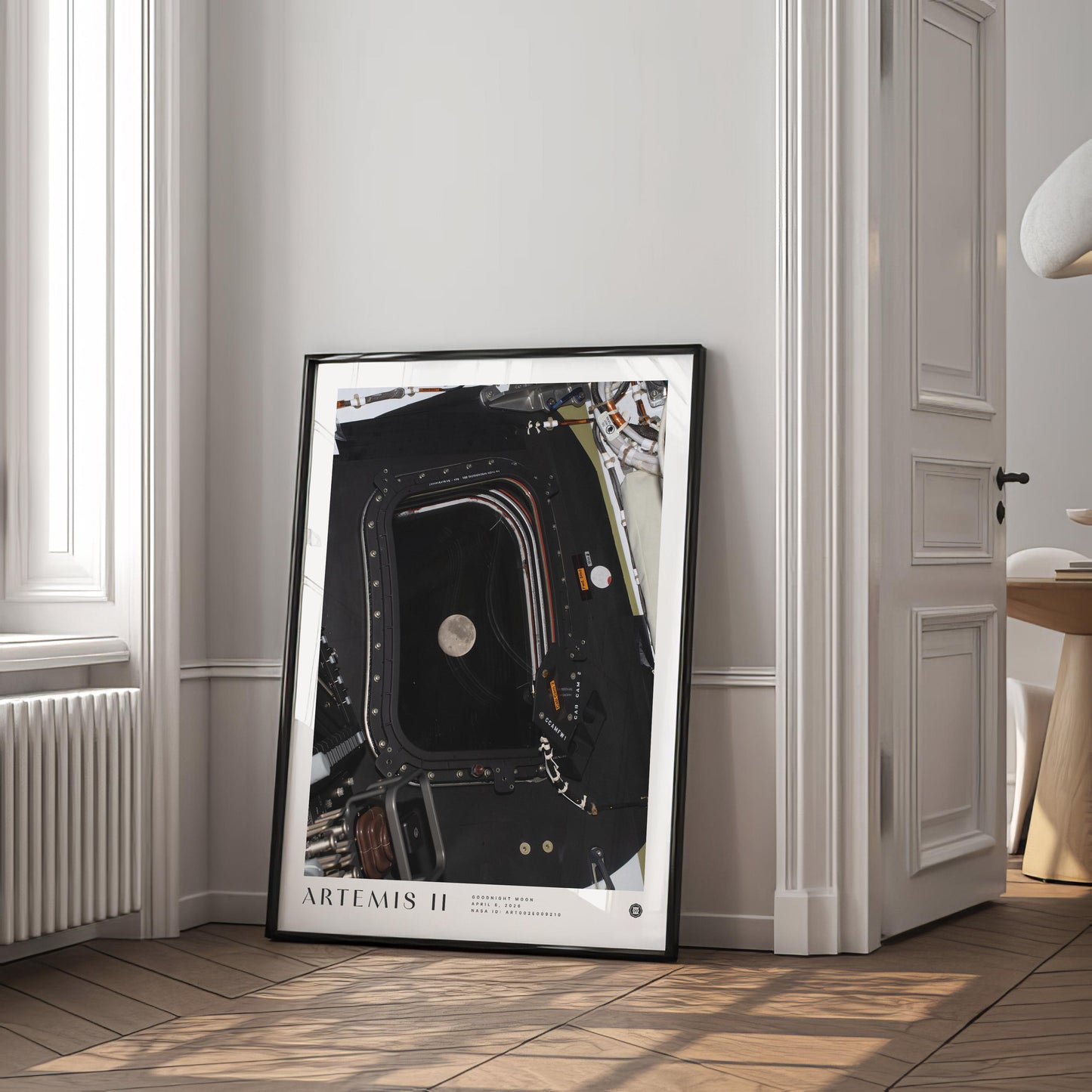 Black-framed Artemis II Goodnight Moon art print leaning against a wall in a bright room beside a window and open doorway.