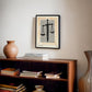 Black-framed Sword & Scales artwork displayed in an interior setting above a wooden bookcase with books and a ceramic vase.
