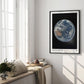 Black-framed Artemis II Earth art print hanging in a sunlit room above a window bench with cushions, books, and flowers.