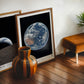 Oak-framed Artemis II Earth art print leaning on a wooden floor beside another space print, a striped wood vase, and a brown stool.