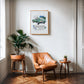 Oak-framed 1961 Jaguar E-Type art print above a tan leather chair and potted plants.