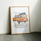Oak-framed orange BMW 2002 artwork leaning against a pale wall between two chairs.