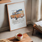 Light wood-framed orange BMW 2002 artwork leaning near a window, books, and a cup of tea.